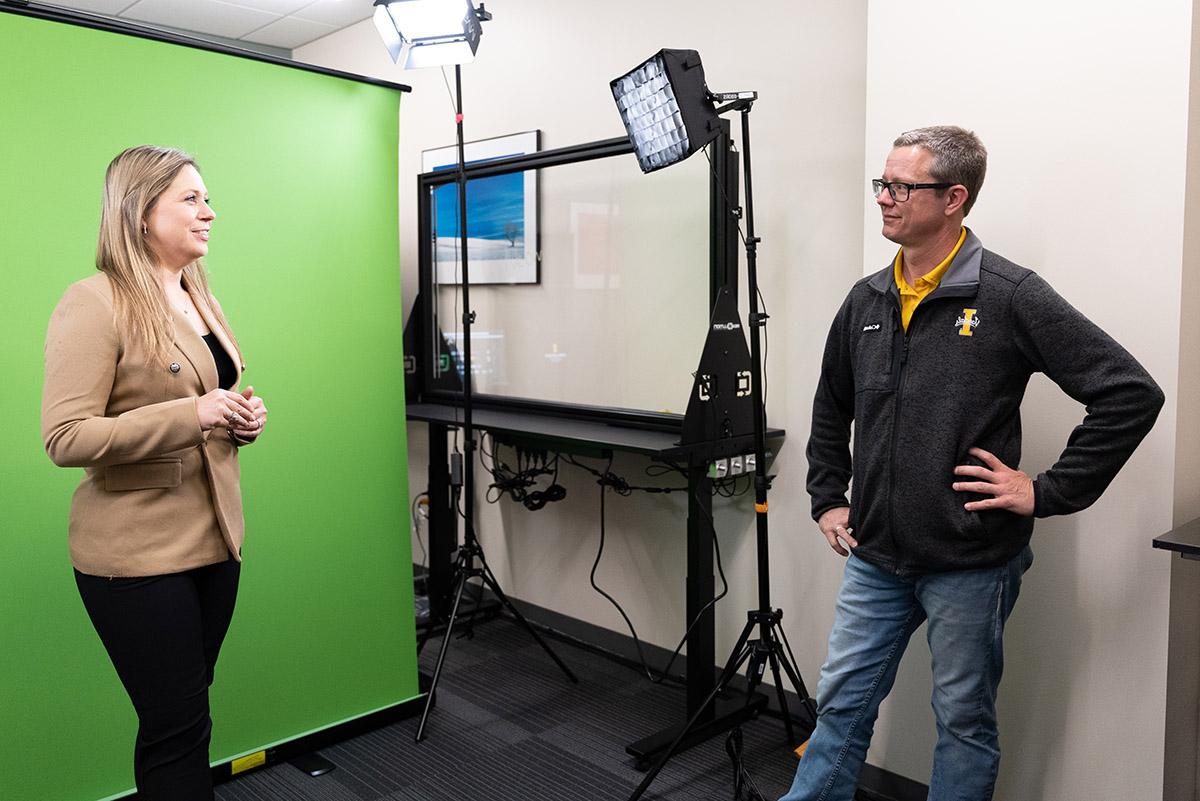 在一个装满录音设备的视频制作工作室里，肖恩·奎伦与站在绿色屏幕前的米亚·格罗扎交谈. 